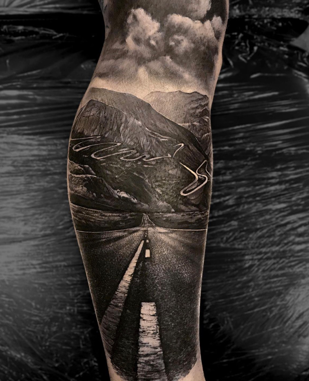 Tattoo-Anansi-Studio-Munchen-Munich-Haidhausen-Alex-leg-landscape-roadtrip-street-highway-asphalt-marks-mountains-nature-best-black-and-grey-realism.jpg